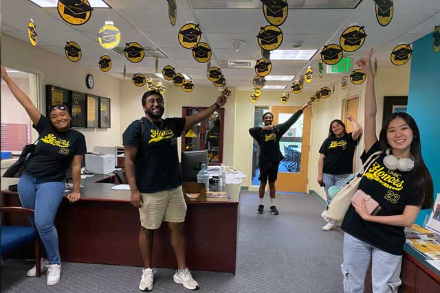 Grads pointing to their graduation hats that are hanging from the ceiling